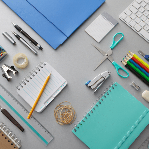 Image showing a group of office supplies on a table. The items include a blue folder with a pen, a keyboard and scissors, and various other objects such as pencils, tape, rubber bands, and spiral notebooks